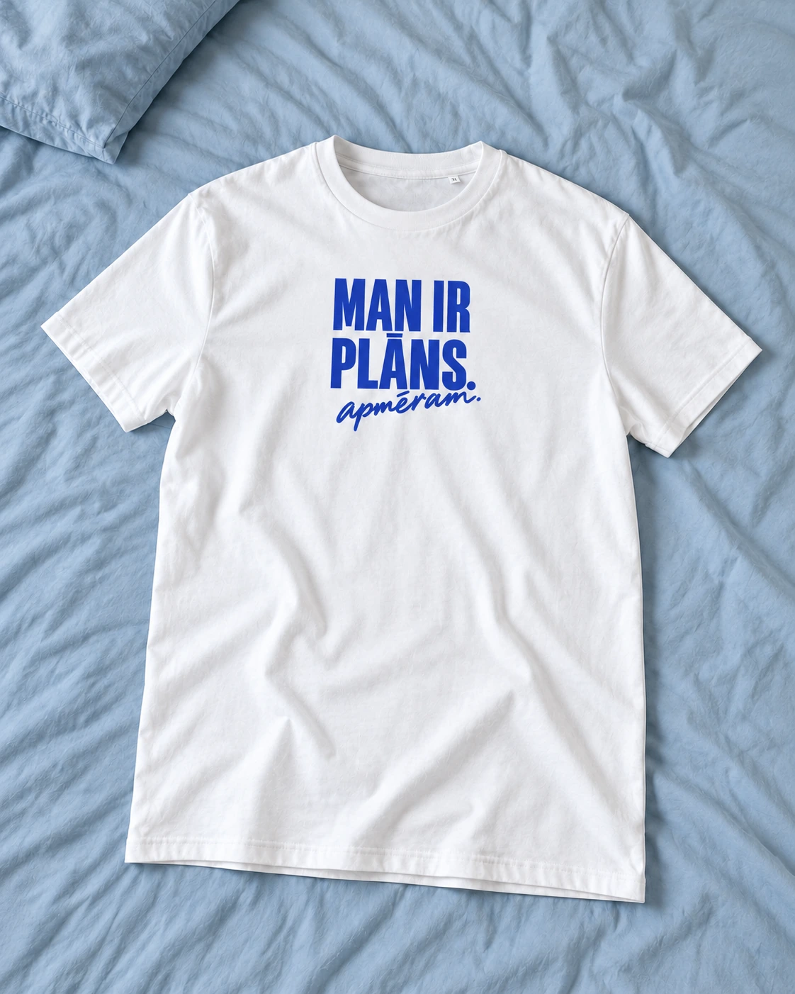 Balts T-krekls “Man ir plāns. Apmēram.” uz gultas